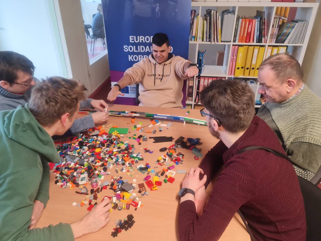 Komandinis užsiėmimas - penki žmonės prie stalo dėlioja tiltą iš lego kaladėlių. Veikla skirta atrasti stiprybes