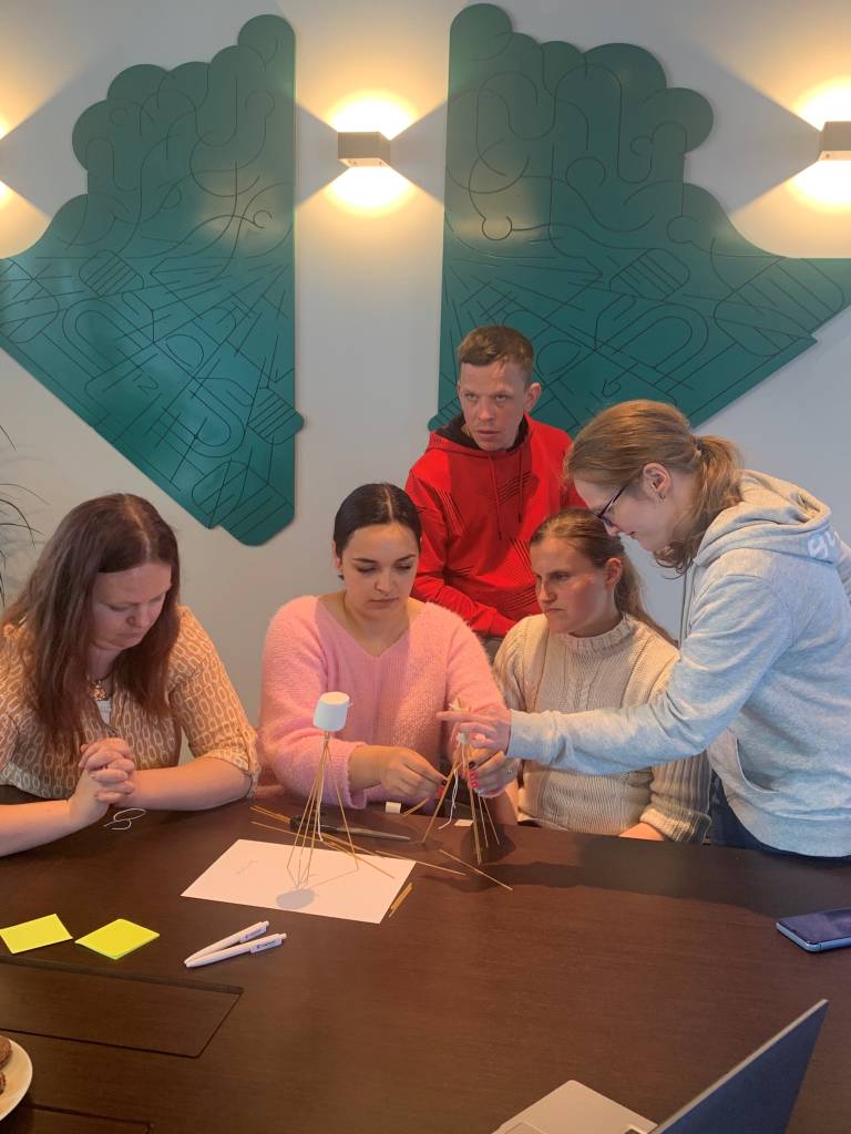 Prie stalo vyksta komandinis užsiėmimas, 5 žmonės kažką konstruoja iš makaronų 